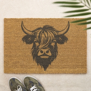 May include: A rectangular coir doormat in shades of brown, featuring a black illustration of a Highland cow's head. The cow has large horns and long, shaggy hair. A pair of camouflage trainers are in the foreground.
