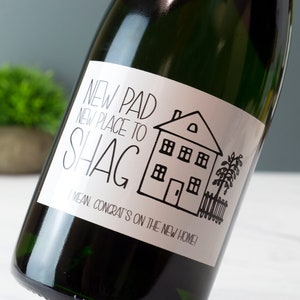 Puede incluir: Una botella de champ&aacute;n con una etiqueta blanca que dice "NEW PAD NEW PLACE TO SHAG I MEAN. CONGRAT'S ON THE NEW HOME!" con una ilustraci&oacute;n en blanco y negro de una casa.