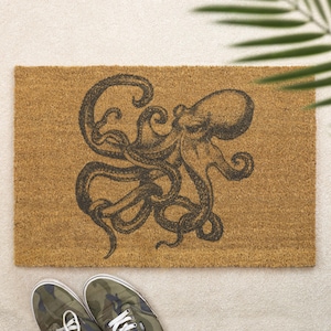 Könnte beinhalten: Eine rechteckige Fußmatte mit einer detaillierten schwarzen Oktopus-Illustration, die auf einem natürlichen braunen Kokosfaser-Hintergrund zentriert ist. Das Oktopus-Design weist komplizierte Linien und Schattierungen auf. Die Matte liegt auf einer hellen Oberfläche, mit Tarn-Sneakers unten.