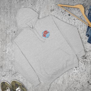 May include: A pale grey hoodie with a front pocket and a hood. The hoodie has a small, colourful emblem on the chest, showing a globe with red and blue details. The hoodie is made of a soft material and is suitable for casual wear.
