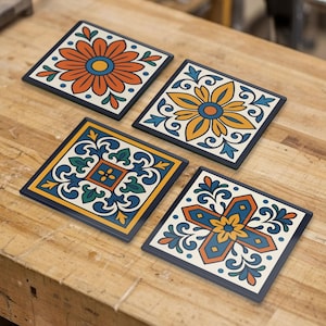 Può includere: Un set di quattro sottobicchieri quadrati con motivi distinti in arancione, giallo, blu e verde. Ogni sottobicchiere presenta un motivo floreale o geometrico unico su uno sfondo color crema con un bordo scuro, disposti su una superficie in legno.