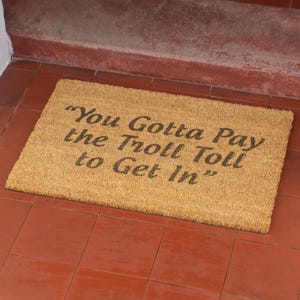 Puede incluir: Una alfombra de coco marrón con el texto "You Gotta Pay the Troll Toll to Get In" en negro.