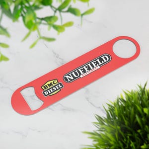 Puede incluir: Un abridor de botellas rojo con el texto "NUFFIELD UNIVERSAL" y "BMC DIESEL" impreso. El abridor tiene un agujero circular en un extremo y un abridor de botellas en el otro.