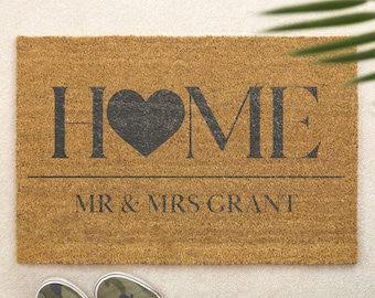 Puede incluir: Una felpudo rectangular de fibra de coco con la palabra "HOME" en letras grises oscuras, con un corazón reemplazando la "O". Debajo, el texto "MR & MRS GRANT". Zapatillas de camuflaje en la parte inferior.