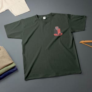 K&ouml;nnte beinhalten: Dunkelgr&uuml;nes T-Shirt mit einer kleinen rot-grauen Grafik auf der Brust. Die Grafik enth&auml;lt das Wort "TRIUMPH". Im Hintergrund sind weitere gefaltete Hemden zu sehen.