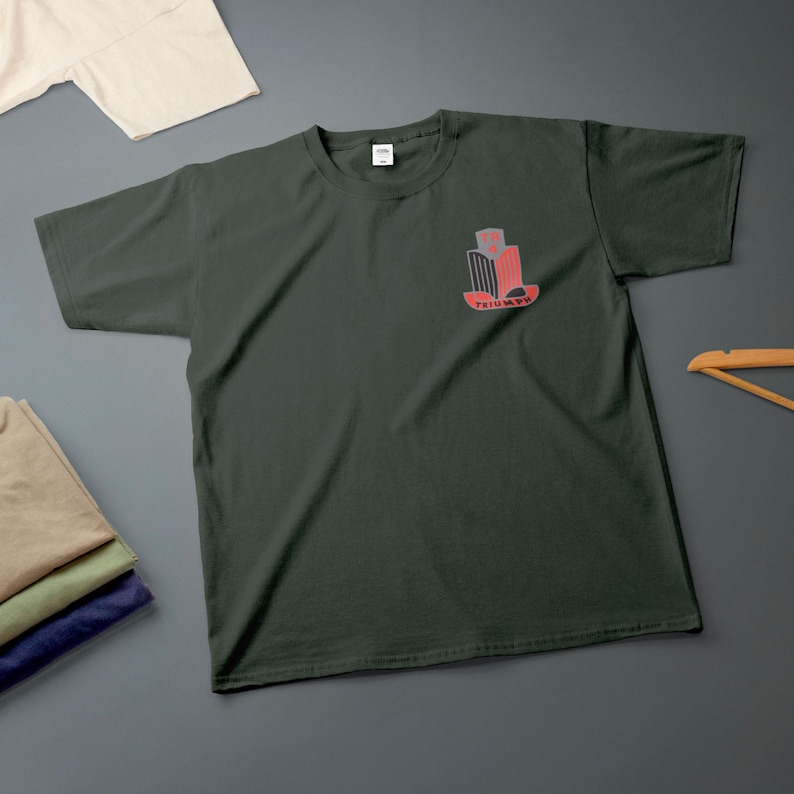 K&ouml;nnte beinhalten: Dunkelgr&uuml;nes T-Shirt mit einer kleinen rot-grauen Grafik auf der Brust. Die Grafik enth&auml;lt das Wort "TRIUMPH". Im Hintergrund sind weitere gefaltete Hemden zu sehen.