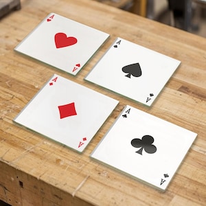 Puede incluir: Un juego de cuatro posavasos cuadrados de cristal que muestran los cuatro ases de una baraja de cartas. Cada posavasos tiene un fondo blanco con un símbolo central grande en rojo o negro, y la letra A impresa en las esquinas opuestas.