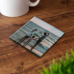 Op de afbeelding: Een vierkante glazen onderzetter met een afgedrukte foto van drie personen die bij zonsondergang in het ondiepe oceaanwater staan. De afbeelding toont een strandscène met zachte golven en een lichte lucht op de achtergrond.