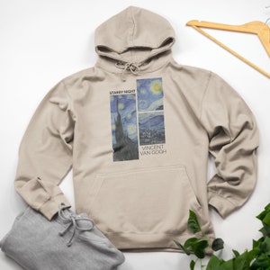 Puede incluir: Una sudadera con capucha beige con una impresión de la pintura "La noche estrellada" de Vincent van Gogh. La impresión incluye el título "La noche estrellada" y el nombre del artista "Vincent van Gogh".