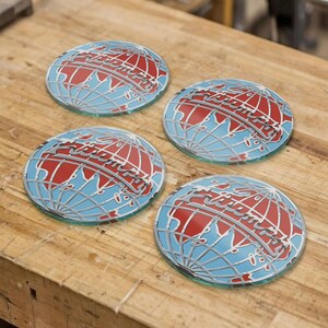 May include: Four round glass coasters with a globe design. The globe features a red landmass, blue oceans, and a silver outline. The word "Liberty" is written across the globe in silver lettering. The coasters are on a wooden surface.
