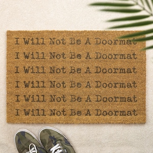 May include: A rectangular doormat made of natural fibres, with the repeated text "I Will Not Be A Doormat" in a dark brown font. The mat is placed on a light-coloured surface, with a pair of camouflage trainers visible at the bottom.