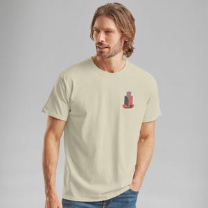 K&ouml;nnte beinhalten: Cremefarbenes T-Shirt mit einem kleinen, stilisierten Geb&auml;ude-Grafik in Rot, Schwarz und Grau. Die Grafik befindet sich auf der linken Brust. Das Shirt hat einen Rundhalsausschnitt und kurze &Auml;rmel.
