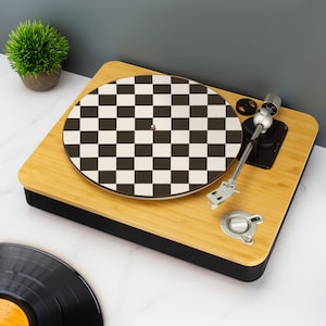 以下が含まれることがあります： 白黒の市松模様のレコードを備えた木製のターンテーブル。 ターンテーブルには、シルバーアームとコントロールノブが付いています。 黒いビニールレコードが前景にあり、鉢植えの小さな緑色の植物が背景にあります。