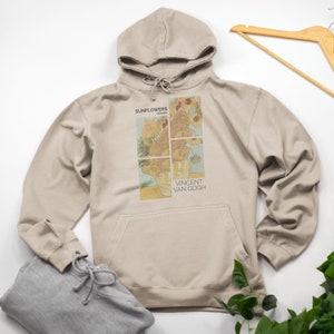 Puede incluir: Una sudadera con capucha beige con una impresión de la pintura "Girasoles" de Vincent van Gogh. La impresión incluye el título "Girasoles" y el nombre del artista "Vincent van Gogh".
