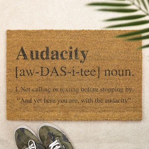 Könnte beinhalten: Rechteckige Fußmatte aus Naturfasern mit dem Wort "Audacity" in großen schwarzen Buchstaben sowie einer Definition und einem Zitat. Tarnfarbene Turnschuhe sind unten im Bild zu sehen.