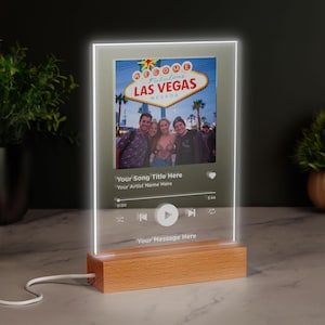 May include: A personalised LED light with a photo and text, set on a wooden base. The image displays a photo with the text "Welcome to Las Vegas Nevada" and music player controls. The text "Your Song Title Here" and "Your Artist Name Here" are also visible.