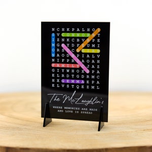 May include: Black acrylic sign with a word search puzzle featuring the words "The McLaughlin's", "Family", "Connor", "Simon", "Gemma", and "Nice". The sign has a white stand and the text "Where memories are made and love is spread" is printed below the puzzle.