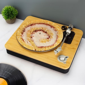 Op de afbeelding: Een bamboe platenspeler met een plaat met cakethema. De plaat heeft een spiraalvormig ontwerp van gele cake, witte glazuur en rode jam. De platenspeler heeft een zwarte basis en zilveren componenten. Een plant in een pot en een vinylplaat zijn ook zichtbaar.