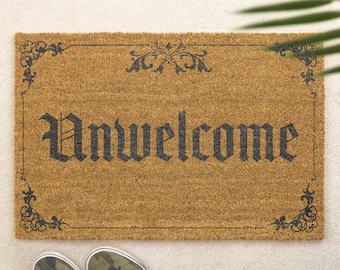 May include: A rectangular doormat made of natural coir fiber, featuring the word "Unwelcome" in a gothic font. The mat has a decorative border with floral scrollwork in black. Camouflage sneakers are at the bottom.
