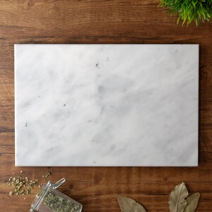 Puede incluir: Una tabla de servir de m&aacute;rmol blanco con una superficie lisa y pulida. La tabla es rectangular y tiene un patr&oacute;n de vetas gris claro.