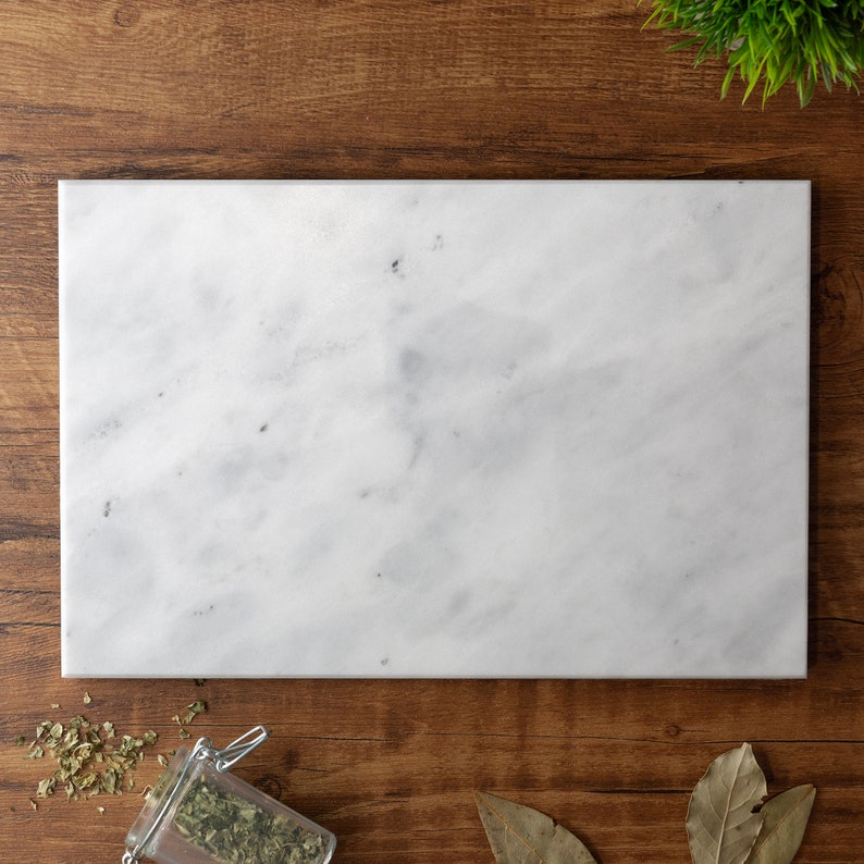 Puede incluir: Una tabla de servir de m&aacute;rmol blanco con una superficie lisa y pulida. La tabla es rectangular y tiene un patr&oacute;n de vetas gris claro.