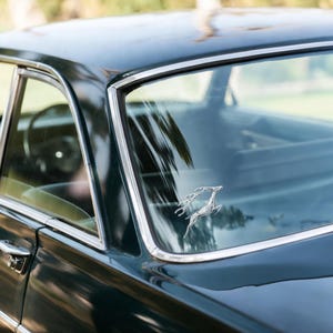 May include: A close-up of a dark green vintage car, showcasing the rear window and a chrome emblem of two leaping deer. The car's body reflects the surrounding environment, with a focus on the classic design.