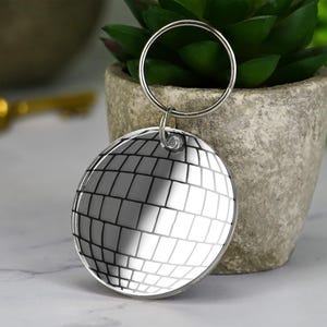 Może przedstawiać: Okrągły, srebrny brelok do kluczy z motywem kuli dyskotekowej. Brelok ma odblaskową powierzchnię z wzorem kratki i srebrny pierścień do mocowania kluczy. W tle mała roślina doniczkowa.