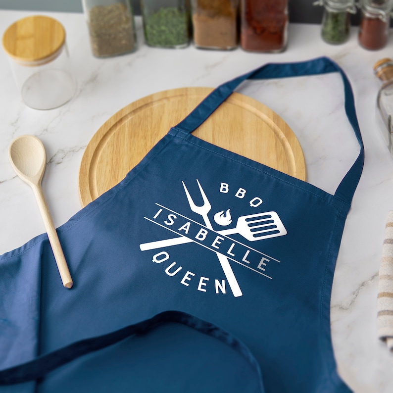May include: A blue BBQ apron with white text that says "BBQ Isabelle Queen" and a white graphic of crossed grilling tools.