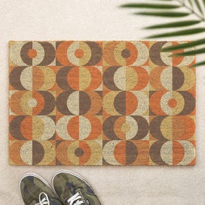 Op de afbeelding: Rechthoekige deurmat met een geometrisch patroon van cirkels en halve cirkels in bruin, oranje en beige tinten. De mat is gemaakt van een textuurrijk materiaal. Een paar camouflage sneakers staan onderaan de afbeelding.