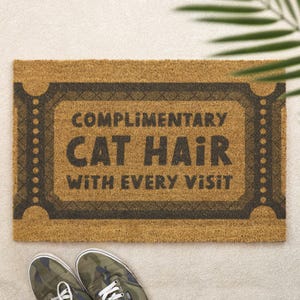 以下が含まれることがあります： 長方形の天然繊維製の玄関マット。テキスト「COMPLIMENTARY CAT HAIR WITH EVERY VISIT」が濃いグレーの文字で書かれています。マットには装飾的なボーダーがあり、明るい色の表面に置かれています。迷彩柄のスニーカーが下部にあります。