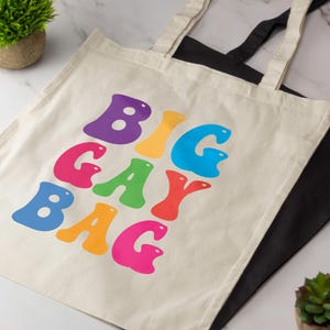 Op de afbeelding: Een crèmekleurige canvas tas met twee schouderbanden, voorzien van de tekst BIG GAY BAG in grote, bubbelachtige, veelkleurige letters in paars, geel, blauw, roze, groen en rood. Een zwarte tas ligt eronder.