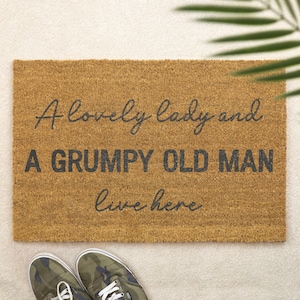 Può includere: Un zerbino rettangolare in fibra di cocco naturale, con il testo "A lovely lady and A GRUMPY OLD MAN live here" in nero. In basso, scarpe da ginnastica con stampa mimetica.