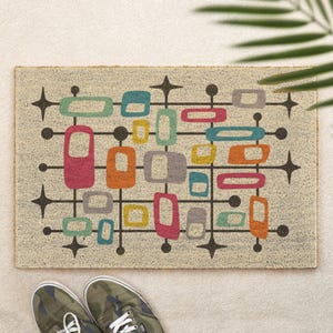 May include: A rectangular doormat with a beige textured surface featuring a mid-century modern pattern of black grid lines, dark circles, and four-pointed stars. The design includes scattered rounded rectangular shapes in teal, pink, orange, yellow, and grey.