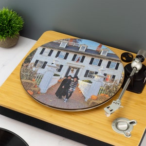 May include: A round wooden record with a photo of a white house with two people in front of it. The house has black shutters and a red door. There are pumpkins in front of the house. The record is on a turntable.
