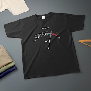 Puede incluir: Una camiseta negra de manga corta con un gráfico blanco de un medidor VU en el pecho. El gráfico incluye el texto "dB level" y "VU" con una aguja apuntando hacia la marca de cero y una zona roja que indica los niveles uno, dos y tres.