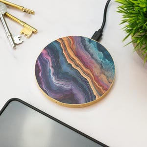 May include: Circular wireless charger featuring a vibrant agate pattern in blue, purple, orange, and gold hues. It has a wooden border and a black charging cable. A smartphone, keys, and a small plant are also visible.