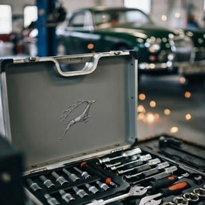 May include: A gray metal toolbox with a silver deer emblem, open to reveal a set of tools. The tools include sockets, wrenches, and screwdrivers. A classic green car is in the background.