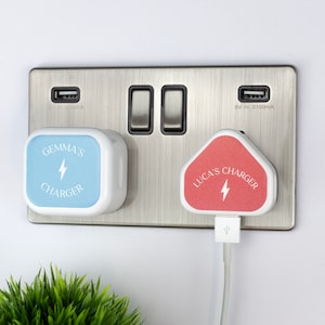 Op de afbeelding: Twee witte USB-wandladers met gepersonaliseerde labels. De blauwe lader zegt "Gemma's Charger" en de roze lader zegt "Luca's Charger". Beide laders hebben een bliksem symbool.