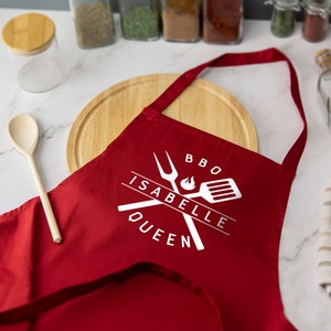 May include: Red BBQ apron with white text that says "BBQ Isabelle Queen" and a graphic of crossed grilling tools with a flame.