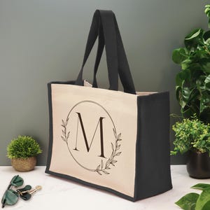 Könnte beinhalten: Eine cremefarbene Canvas-Tragetasche mit schwarzem Besatz und Griffen. Die Tasche hat ein schwarz-weißes Design mit einem Kreis, einem Blattkranz und dem Buchstaben "M" in der Mitte.