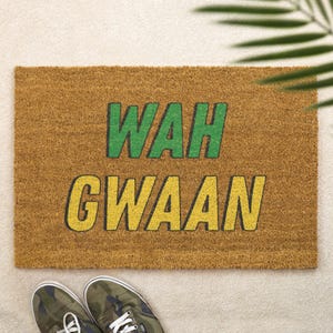 Op de afbeelding: Een rechthoekige deurmat van natuurlijk bruin kokosvezel. De mat heeft de woorden "WAH GWAAN" in groene en gele blokletters. Onderin de mat zijn camouflage sneakers te zien.