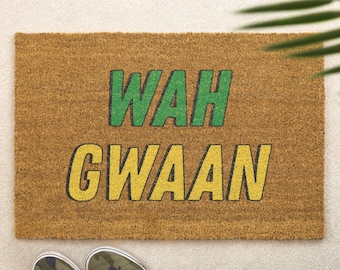 Pode incluir: Um capacho retangular feito de fibra de coco marrom natural. O tapete apresenta as palavras "WAH GWAAN" em letras de bloco verdes e amarelas. Um par de tênis camuflados estão na parte inferior do tapete.