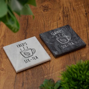 May include: Two square coasters featuring a teacup design. One is white with "Fred's Six-Tea" text, the other is dark grey with "Claire's Six-Tea" text. The coasters are displayed on a wooden surface.