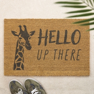 May include: A rectangular doormat made of natural coir fiber. The mat features a black giraffe graphic and the words "HELLO UP THERE" in a handwritten style font. A pair of camouflage sneakers are at the bottom of the image.