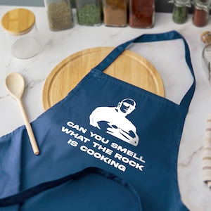 May include: A navy blue apron with white text and a graphic of a bald man. The text reads "CAN YOU SMELL WHAT THE ROCK IS COOKING". A wooden spoon and cutting board are in the background.