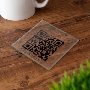 May include: A square glass coaster featuring a black QR code printed in the center. The text "WELCOME TO" is printed above the code, and the text "SCAN TO JOIN OUR WI-FI" is printed below the code in a simple, dark, sans-serif font.