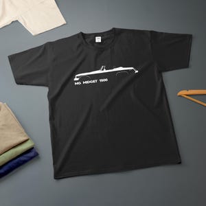 Może przedstawiać: Czarny t-shirt z białym obrysem kabrioletu MG Midget 1500. Tekst "MG Midget 1500" jest wydrukowany pod sylwetką samochodu. Koszulka jest prezentowana na szarej powierzchni z innymi złożonymi koszulkami i drewnianym wieszakiem.
