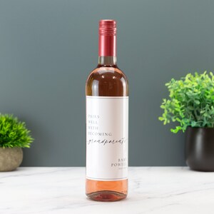 May include: A bottle of rose wine with a white label that says "Pairs well with becoming grandparents" and "Baby Powell" in black text.