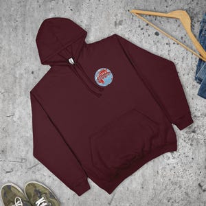 May include: A maroon hooded sweatshirt with a front pocket and a hood. The sweatshirt has a small embroidered globe design with red and blue detailing on the upper left chest. The garment is displayed on a grey surface.