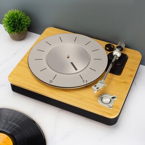 May include: A contemporary turntable with a light wood grain finish and a silver record player. The turntable features a silver dial with black markings and a black base. A vinyl record is visible in the lower left corner.